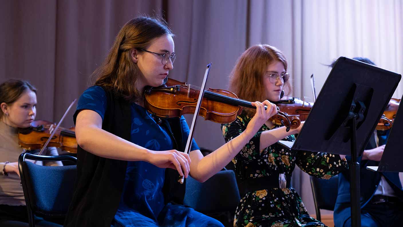 På bilden syns tre personer som sitter på en scen och spelar violin, framför var sitt notställ. De ser koncentrerade ut och fokuserar på sina noter.