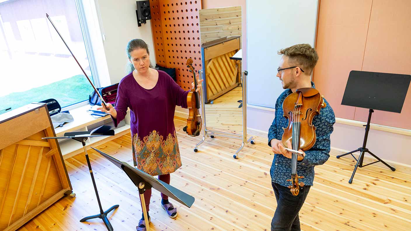 Två personer står med violiner i händerna i ett ljust och öppet övningsrum med trägolv, där det även finns ett piano. Personen till vänster gestikulerar och visar något för personen till höger, som avslappnat håller sin violin.