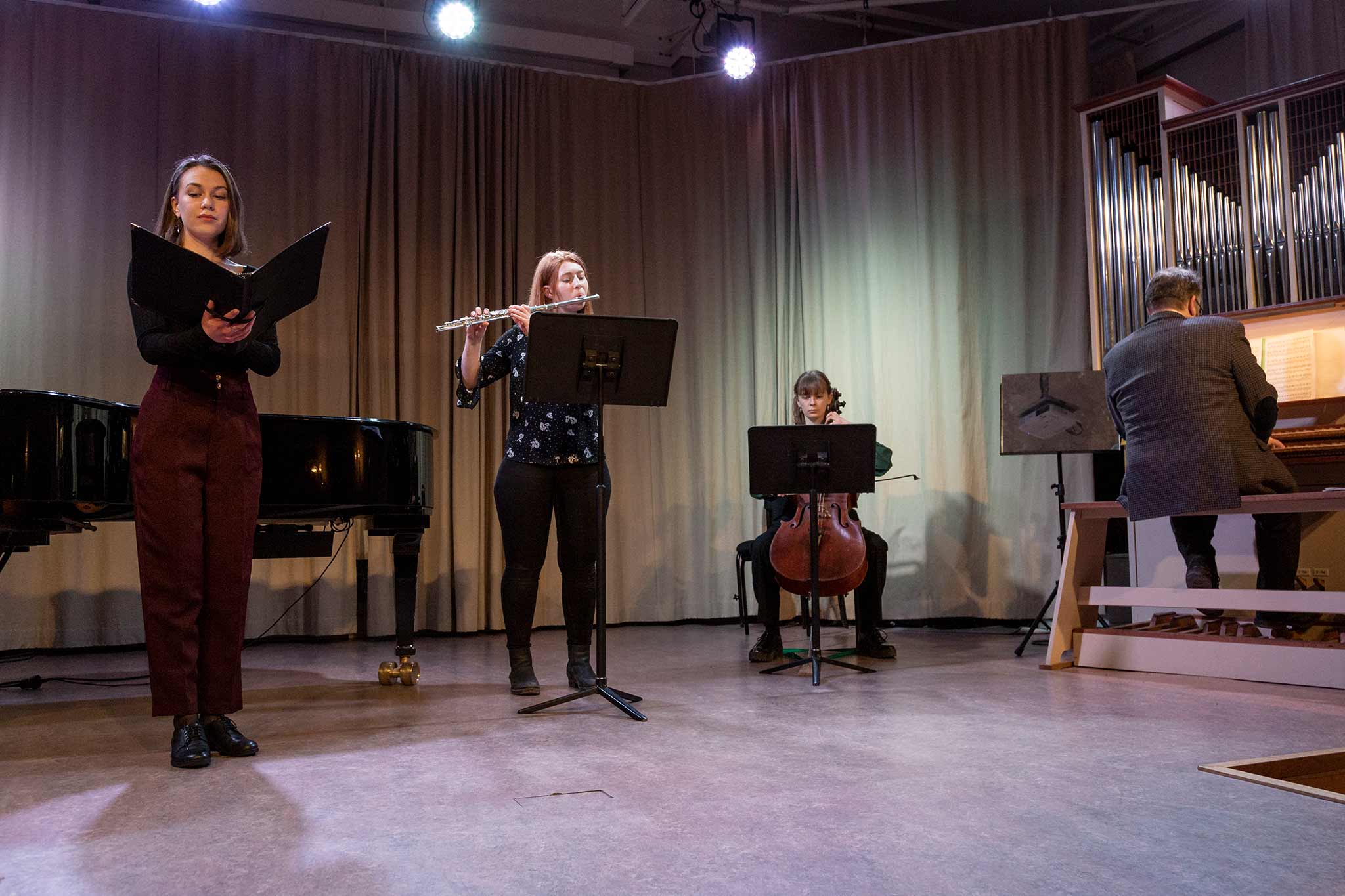 En klassisk ensemble med sångare, flöjtist, cellist och organist framträder på en enkelt upplyst cen. 