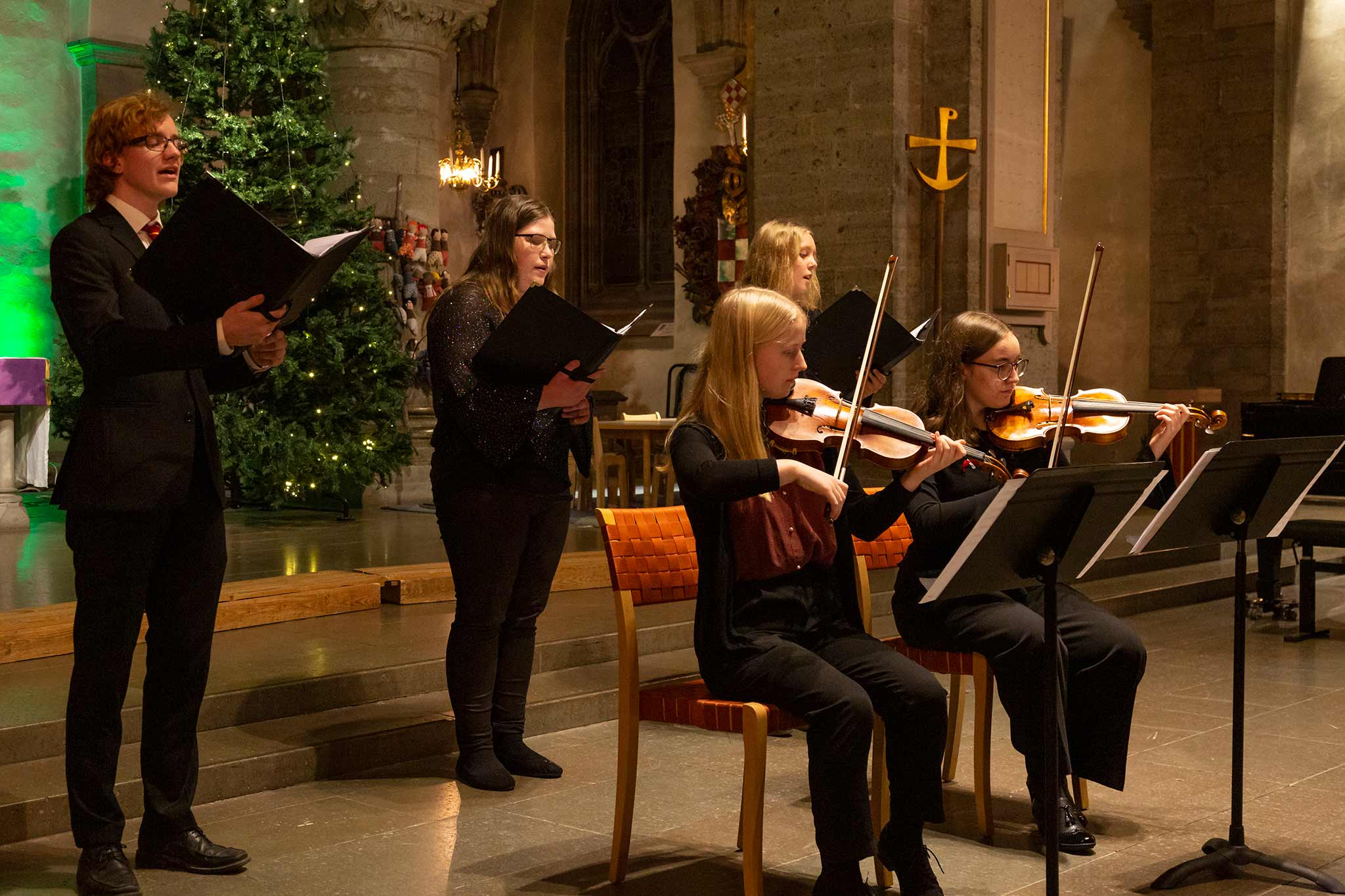 En kammarmusikensemble spelar i en kyrka. Ensemblen består av tre sångare som står bakom två sittande violinister.