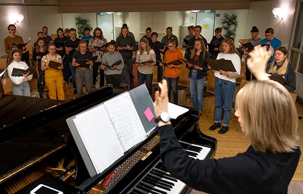 På bilden syns en körövning. En grupp människor står på golvet i en stor aula, håller i notblad och sjunger. I förgrunden dirigerar en person kören med ryggen mot kameran, och framför dirigenten finns en öppen flygel med noter på. 