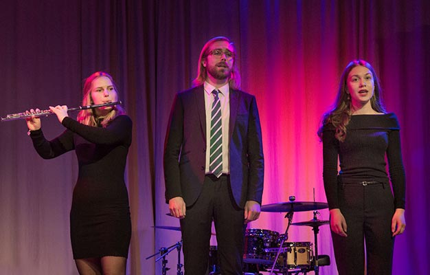 Tre musiker - en som spelar fiol och två som sjunger - framträder på en scen med lila bakgrundsbelysning. Alla bär mörka formella kläder.