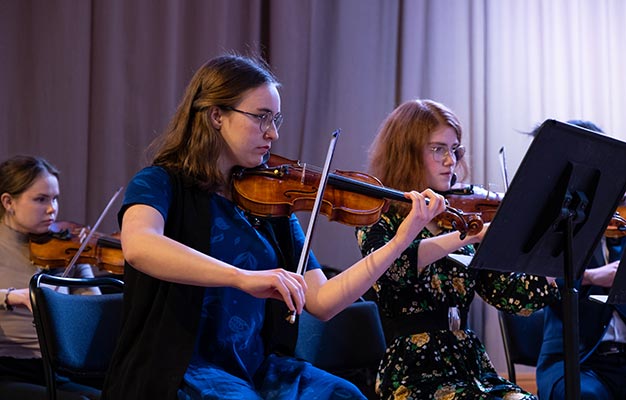 På bilden syns tre personer som sitter på en scen och spelar violin, framför var sitt notställ. De ser koncentrerade ut och fokuserar på sina noter.