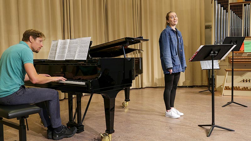 Två personer övar på en scen som är upplyst med vanligt ljus; ingen scenbelysning är igång. Till vänster sitter en person vid ett svart piano. Till höger står en persor framför ett notställ och sjunger. De bär båda informella vardagskläder.