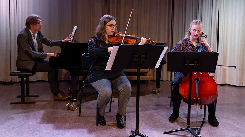 Tre musiker spelar på en scen med skirt lila och blått ljus. Från vänster till höger ser vi en person som spelar piano, en annan som spelar violin och en tredje som spelar cello. Varje musiker har ett notställ framför sig med noter på det. Alla tre är upplädda som för ett scenframträdande.