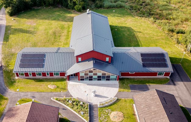 Musikhuset fotograferat från luften. Byggnaden är formad som ett T, med en lång central del och två flyglar. Taket är grått och det finns solceller på de båda flyglarna. Fasaden som syns är röd. Entrén markeras av en hel vägg täckt av fönster och en stenlagd båge i marken. Rund musikhuset, i kanten av bilden, syns delar av flera andra byggnader.