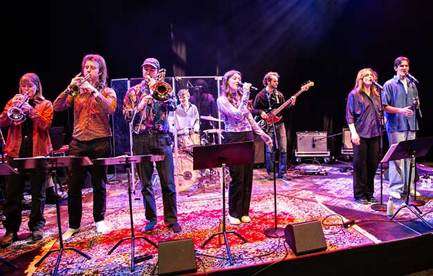 Ett stort band spelar på en scen täckt med orientaliska mattor. Bandet består av tre blåsare, en trummis, en basist, en leadsångare och två sångare som körar.