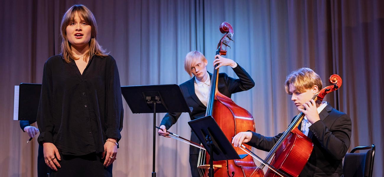 En klassisk ensemble spelar på scen. En sångare i svarta kläder står till vänster i bild. Till höger står en person i vit skjorta och svart kavaj som spelar kontrabas. Snett framför denne sitter en cellist, också klädd i vit skjorta och svart kavaj.