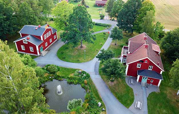 Drönarbild där vi tittar ner på Kävesta folkhögskolas skolområde från ovan. I bild syns två stora röda hus som är belägna mitt emot varandra. Mellan dem går en asfalterad gångväg, som också snirklar sig förbi en damm. Byggnaderna ligger i en lantlig miljö, i fjärran skymtar en åker.