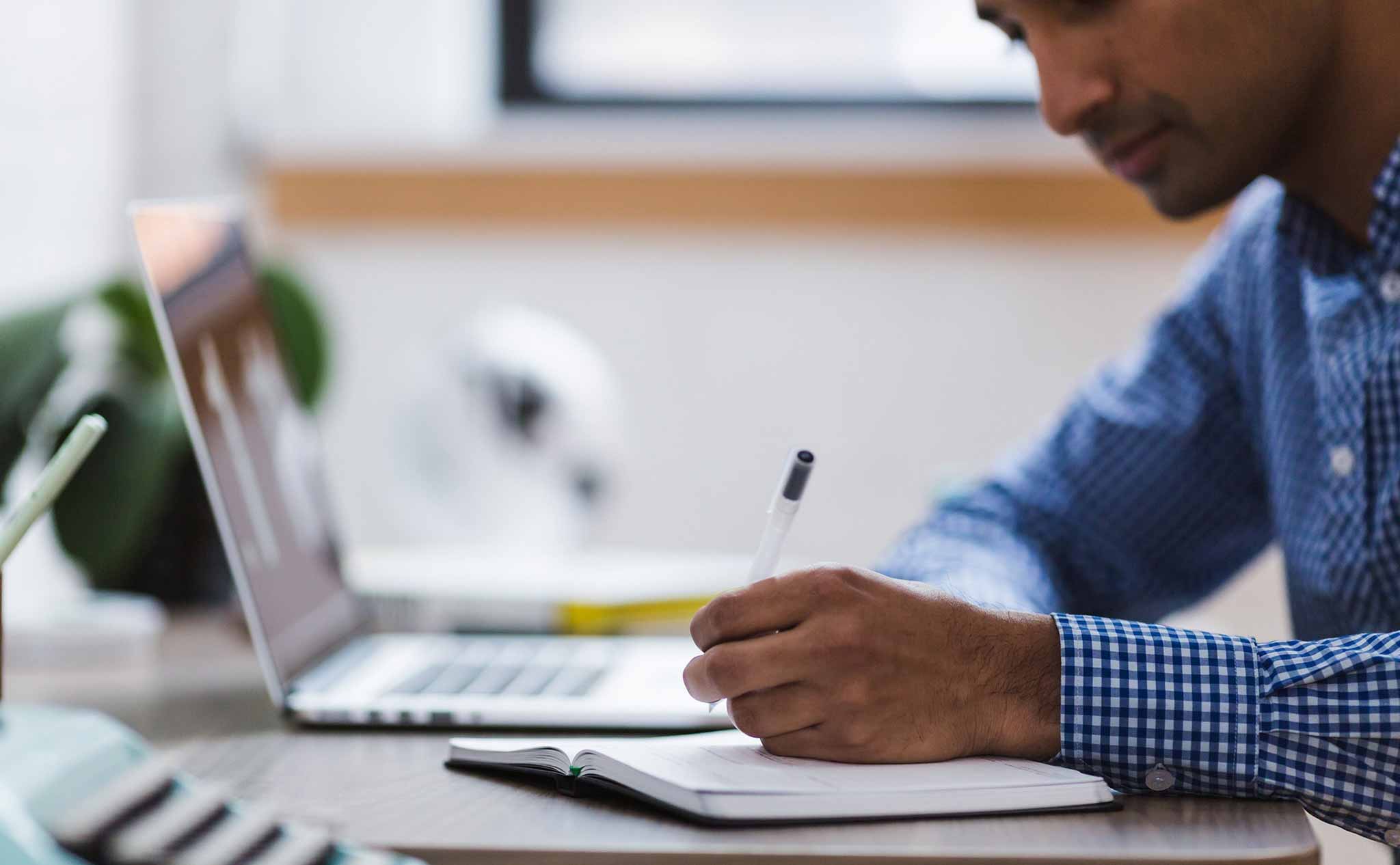 En person med blå- och vitrutig skjorta skriver i ett anteckningsblock och har en bärbar dator öppen framför sig.
